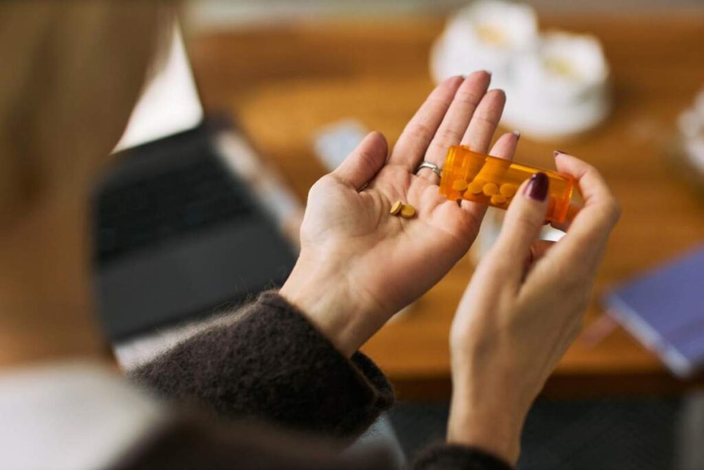 A family member helps an older adult use a pill organizer at a kitchen table, with medication bottles nearby.